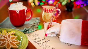 Close up photo of Santa Claus' hand and arm, writing a "nice" list with cookies, milk, lights and a tree in background